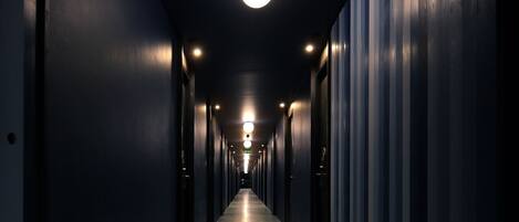 A long, narrow hallway with a speckled floor, flanked by dark walls and doors, leading to a brightly lit end.