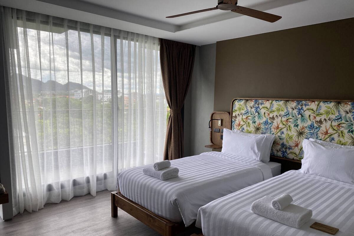 A hotel room with two beds, a wooden headboard, a nightstand, a lamp, and a view through sheer curtains.