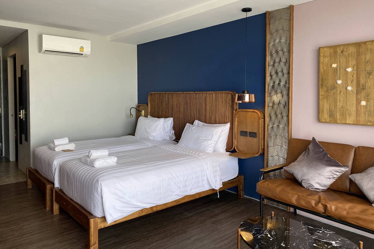 A hotel room with a bed, a wooden headboard, a brown leather sofa, a glass coffee table, and a wall-mounted air conditioning unit.