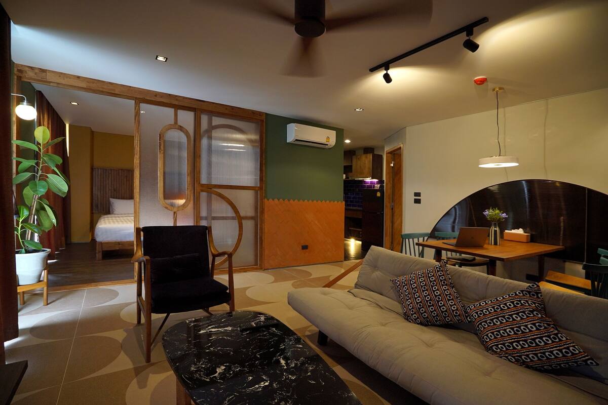 A modern hotel room with a sofa, a black ottoman, a dining table with a laptop, and a potted plant.
