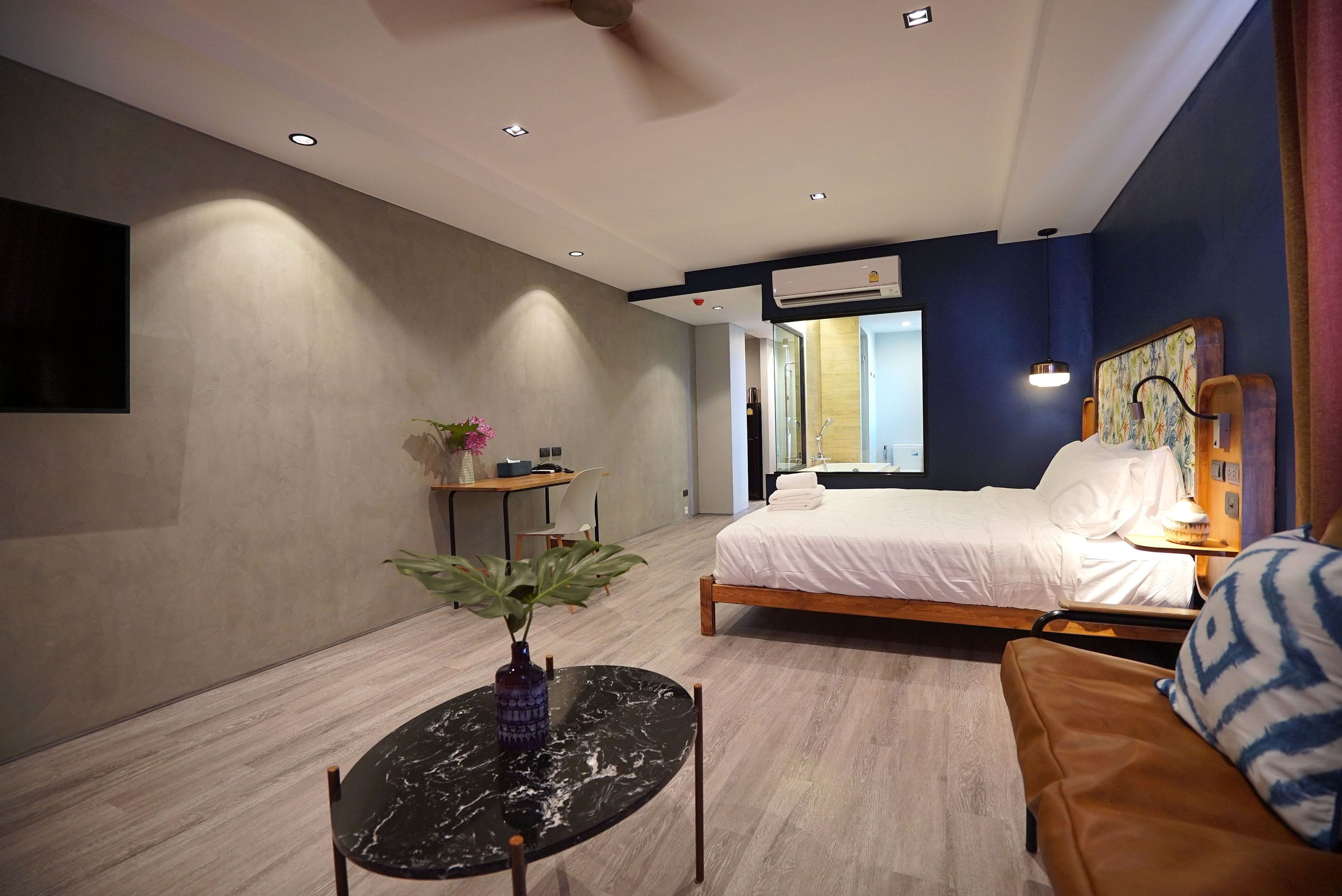 A modern hotel room with a bed, a sitting area with a table and a plant, and a large window offering a view of the outdoors.
