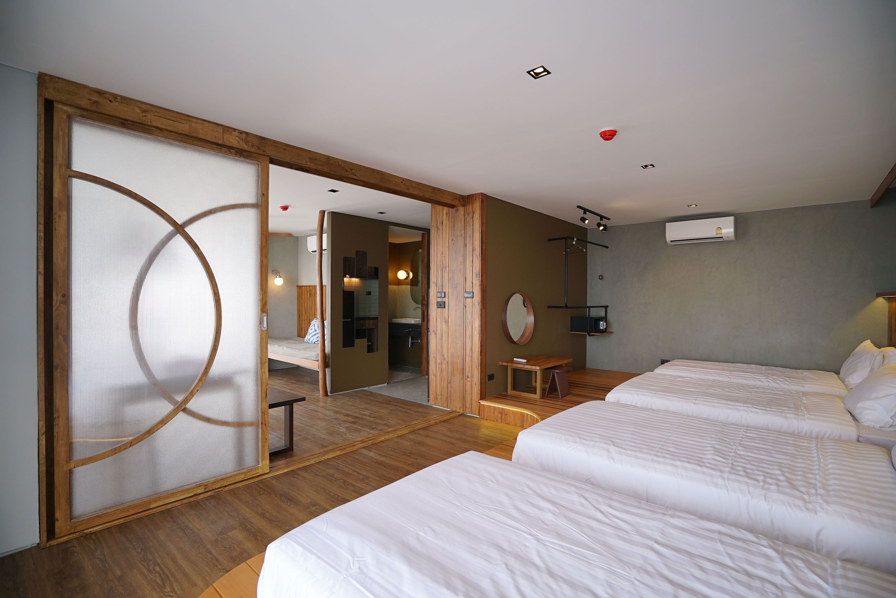 Een hotelkamer met twee bedden, houten vloeren, een schuifdeur met een rond ontwerp, een klein houten krukje en een spiegel aan de muur.