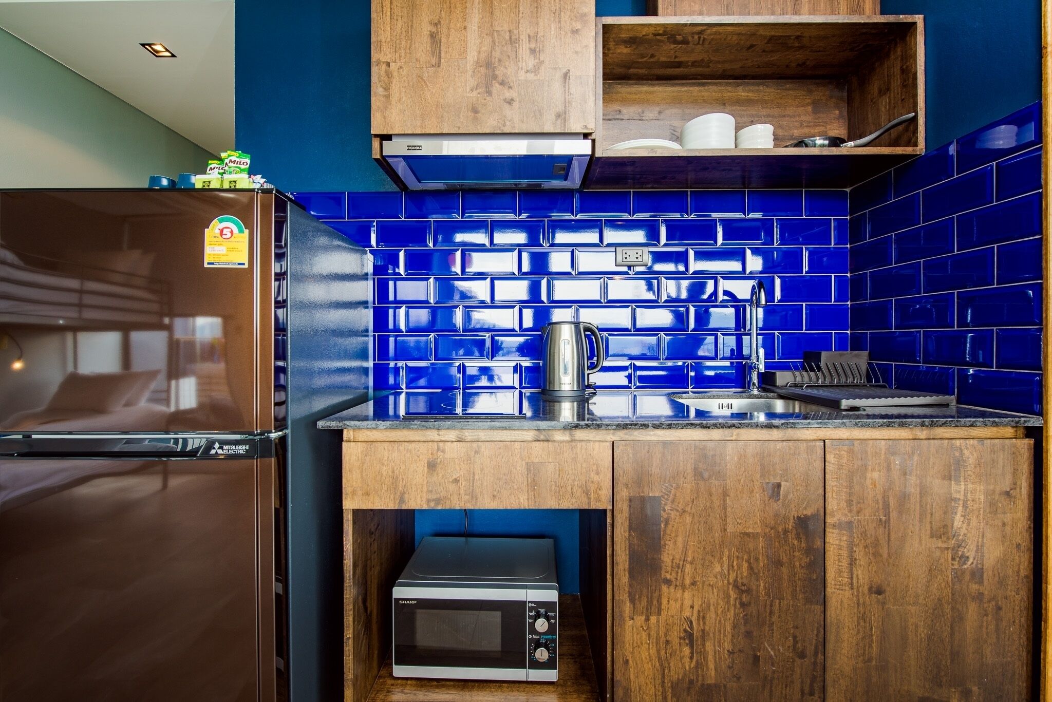 A compact kitchen with blue-tiled backsplash, wooden cabinets, a built-in microwave, and a stainless steel kettle.
