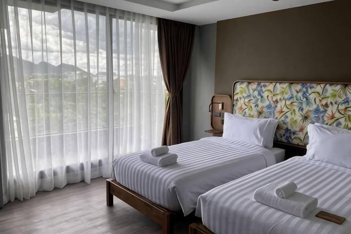 A hotel room with two beds, a large window with sheer curtains, and a view of the outdoors.