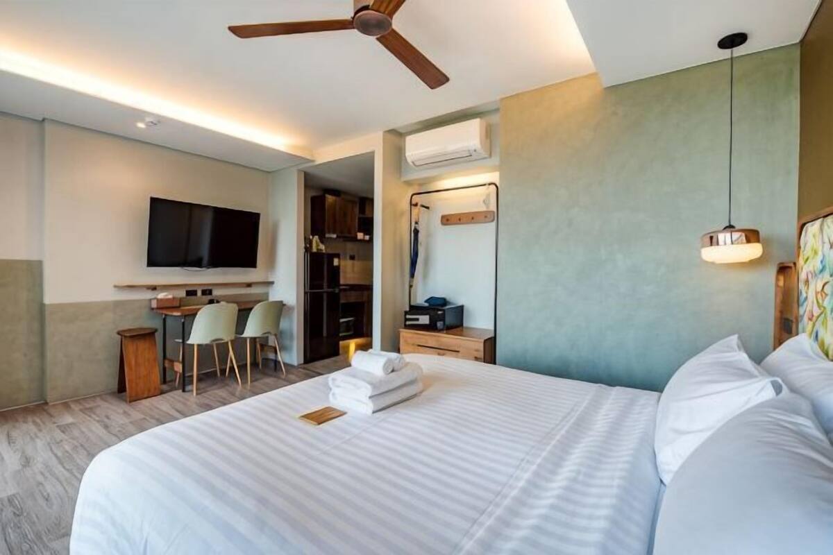 A hotel room with a bed, a ceiling fan, a TV, a desk with a chair, and a mirror.