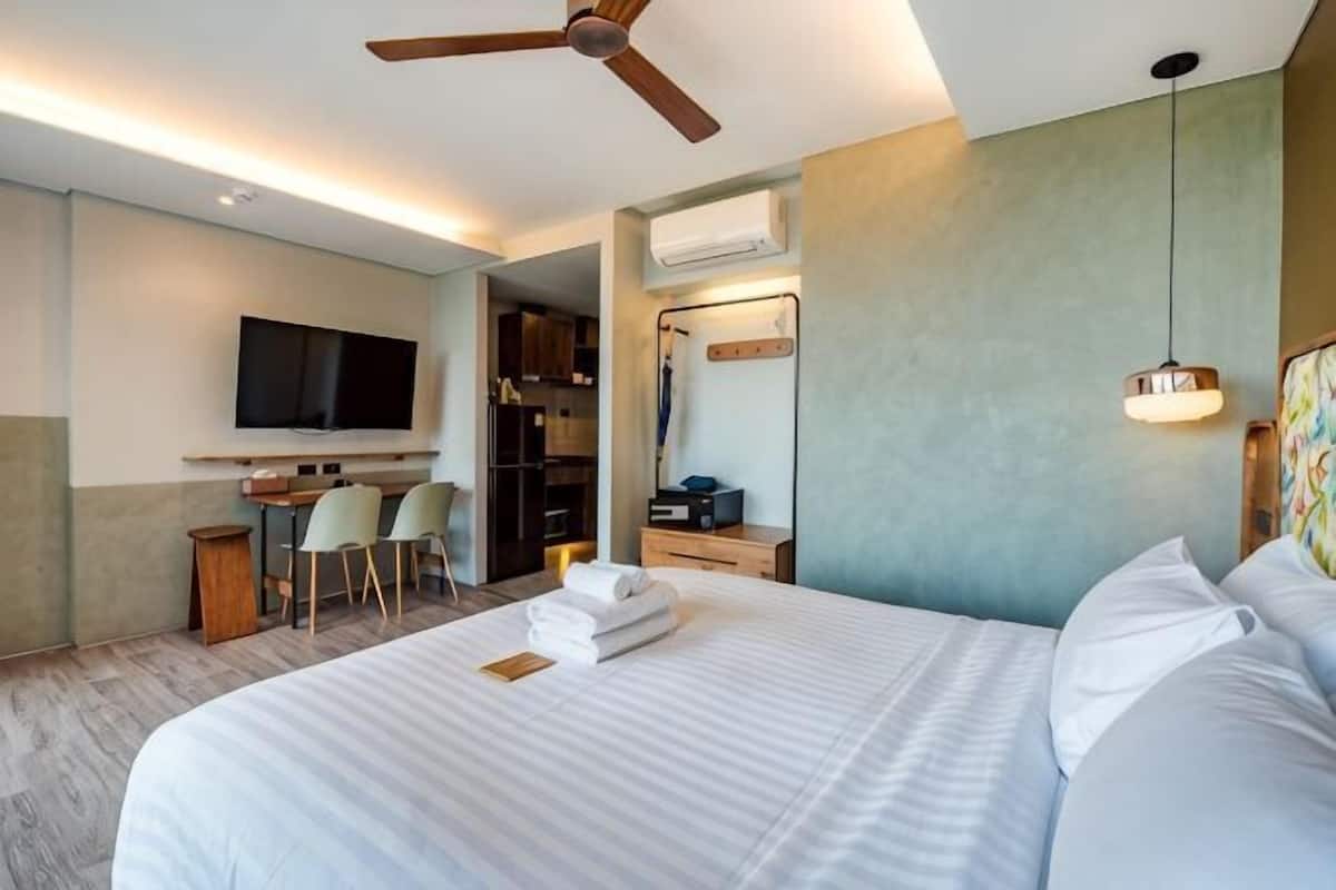 A hotel room with a bed, a ceiling fan, a TV, a desk with a chair, and a mirror.