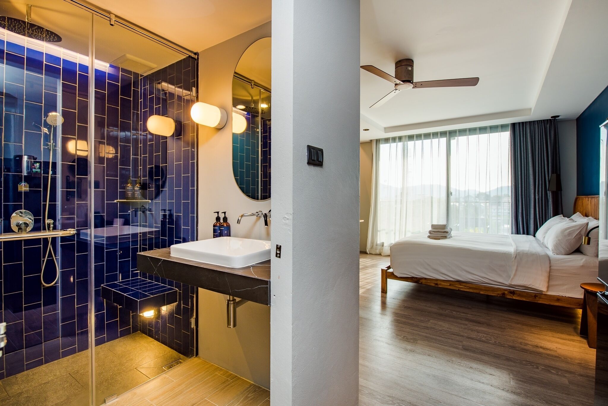 A modern hotel room with a glass-enclosed shower, a large mirror, a wooden bed, and a view of the city through large windows.