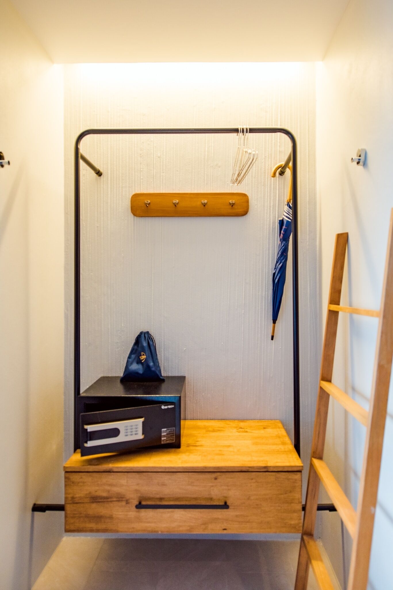 A compact, modern room with a wooden bed, a small bedside table, a drawer, a wall-mounted shelf with hooks, and a ladder.