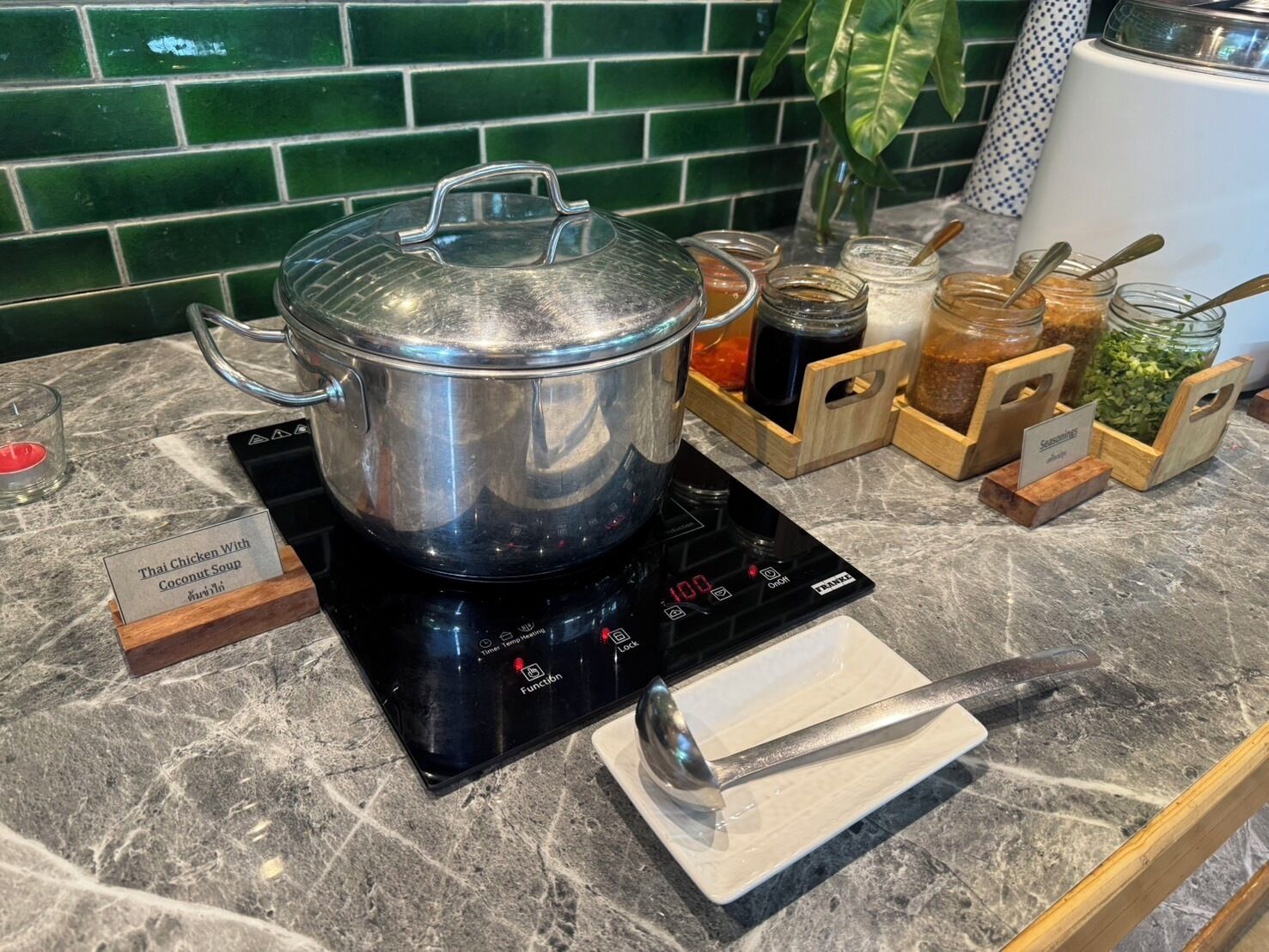 Una cocina con una olla grande sobre una placa de inducción, un vaso de agua y varios condimentos en recipientes de madera.
