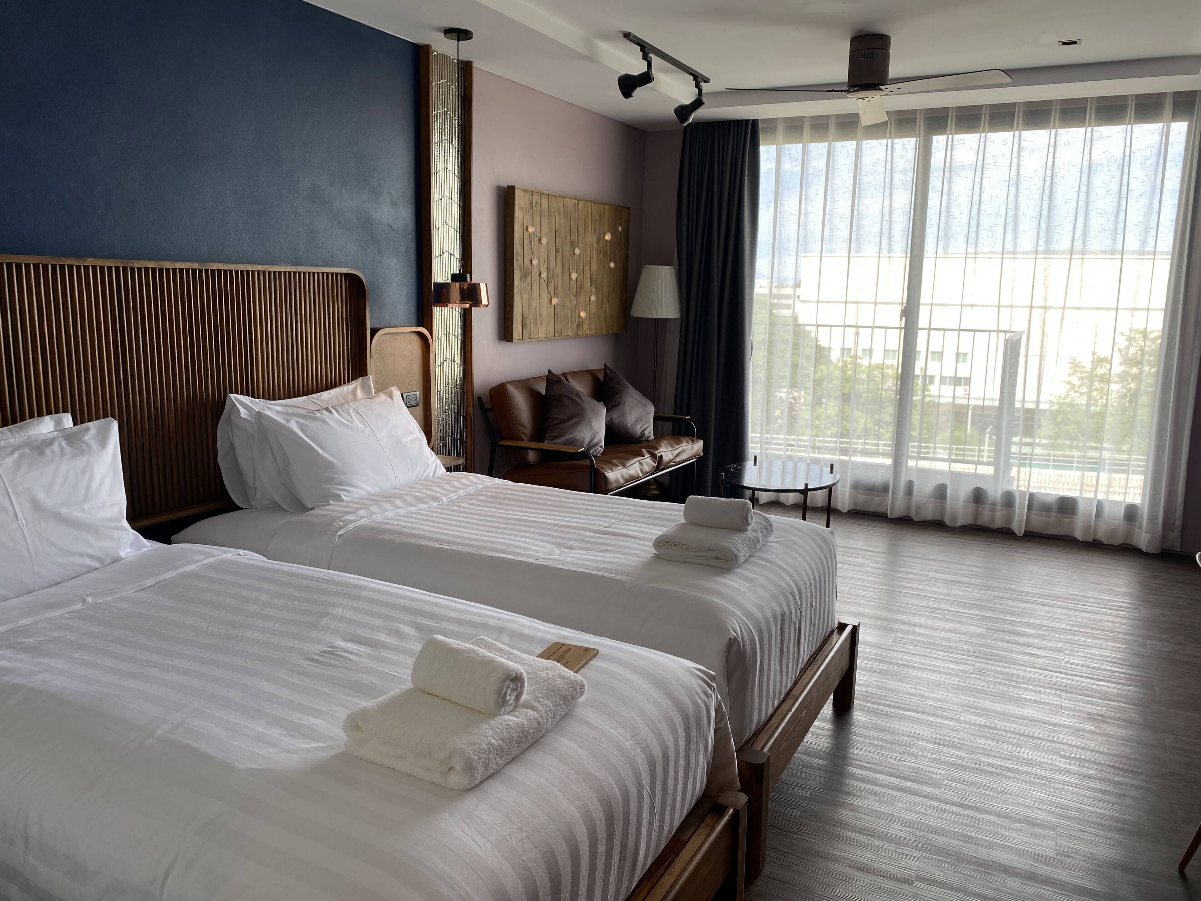 A hotel room with two beds, a wooden headboard, a small table, and a view of the outdoors through large windows.