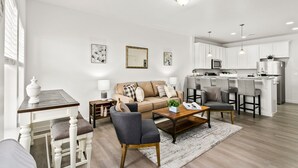 Un salon moderne avec un canapé, une table basse et des fauteuils. On aperçoit, au fond, une cuisine avec des meubles blancs et un coin bar.