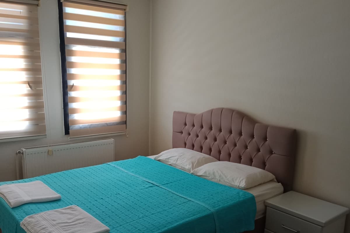 Quarto com cama, mesa de cabeceira, radiador e janela com persianas.