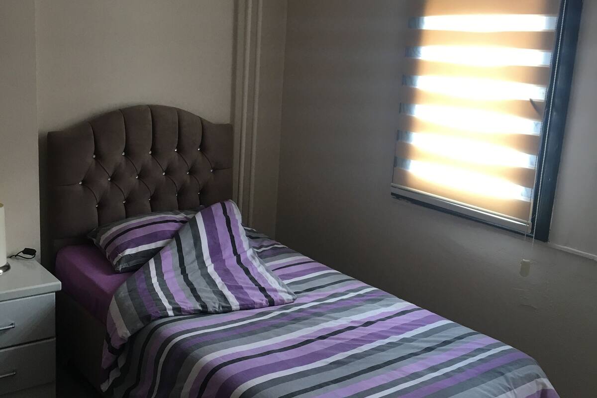 Quarto com uma cama de solteiro, mesa de cabeceira, luminária e janela com persianas.