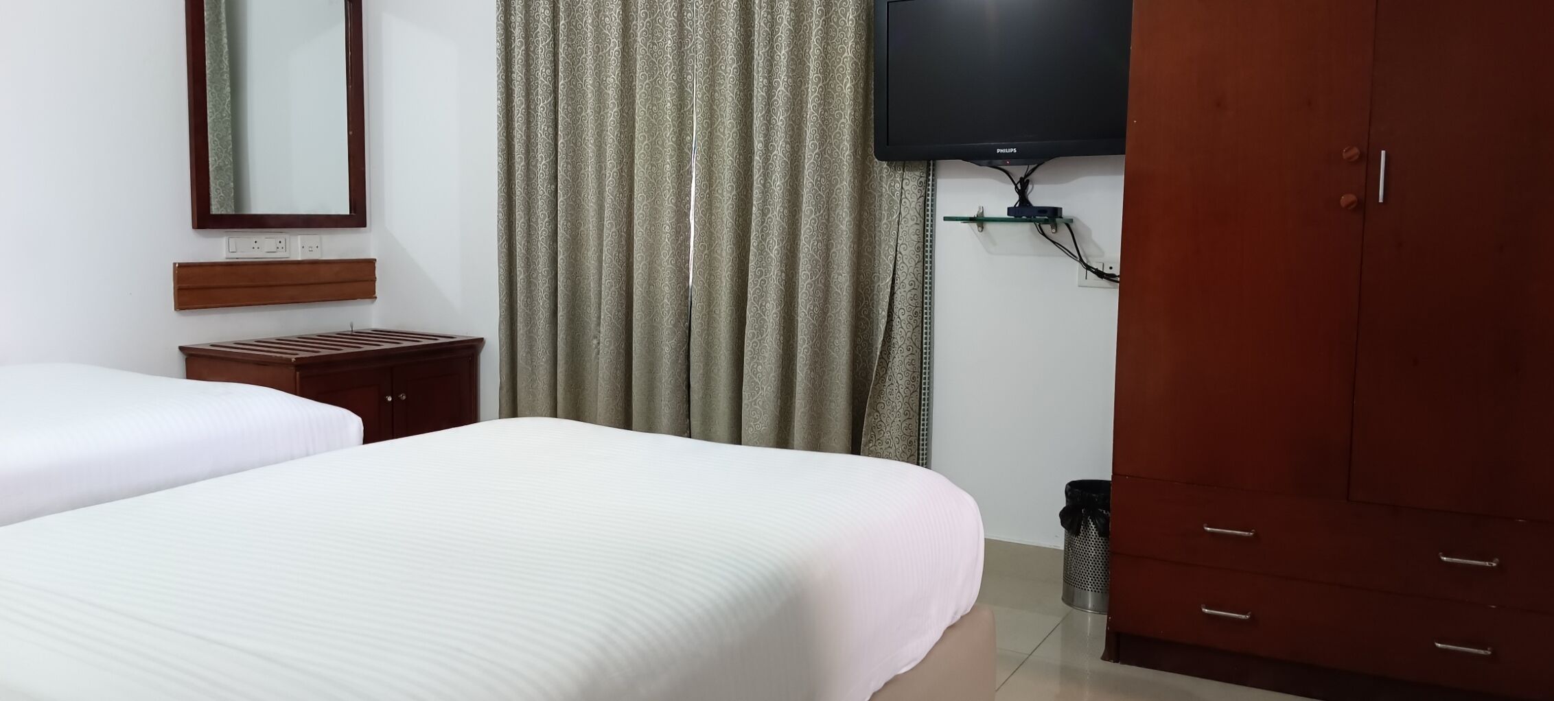 Une chambre d’hôtel avec deux lits, une table de chevet en bois, un téléviseur fixé au mur et une armoire.
