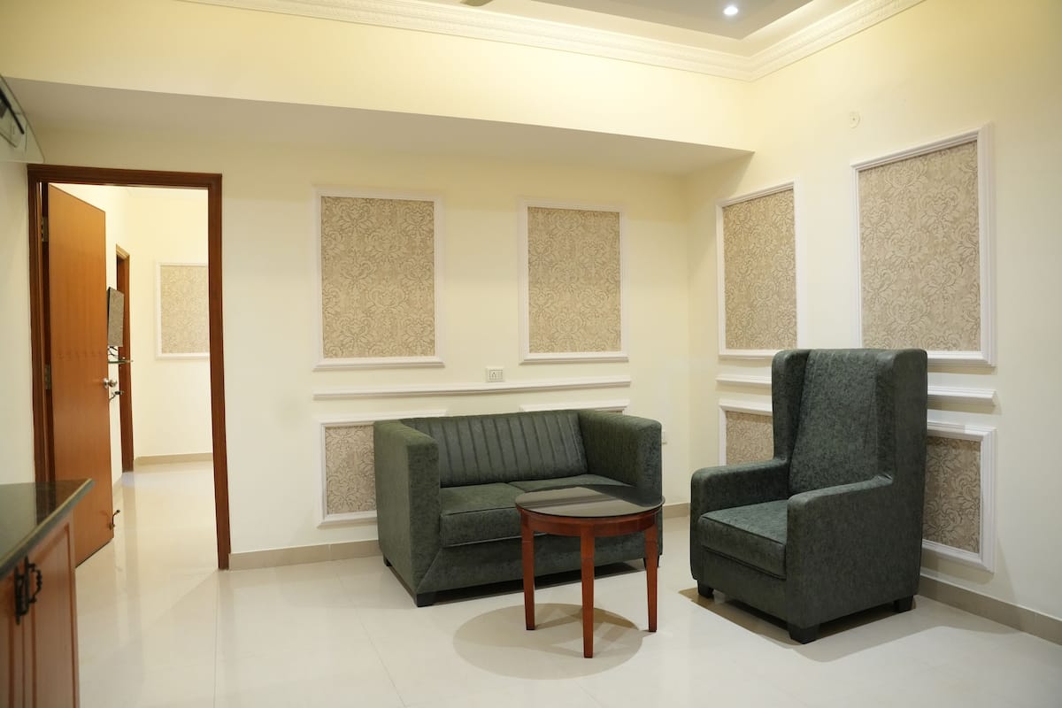Una habitación con un sofá verde, un sillón a juego y una pequeña mesa de madera. Hay paneles decorativos en las paredes y una puerta que da a otra habitación.