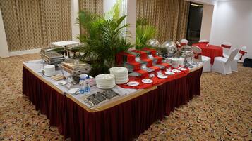 Disposição de buffet com uma longa mesa coberta por uma toalha vermelha, rodeada por cadeiras brancas com fitas vermelhas. Há diversos alimentos e utensílios sobre a mesa, e um grande vaso de planta no centro.