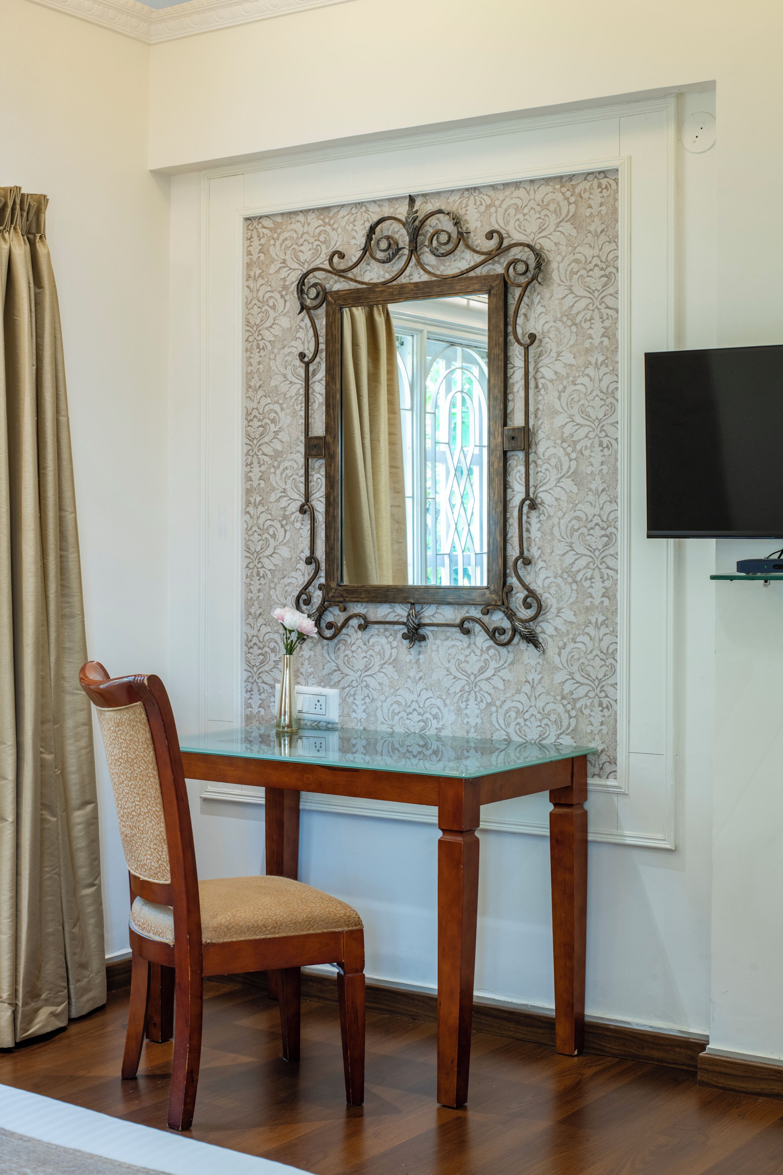 Una habitación con una silla de madera, una mesa de vidrio, un espejo con marco decorativo y un televisor en la pared.