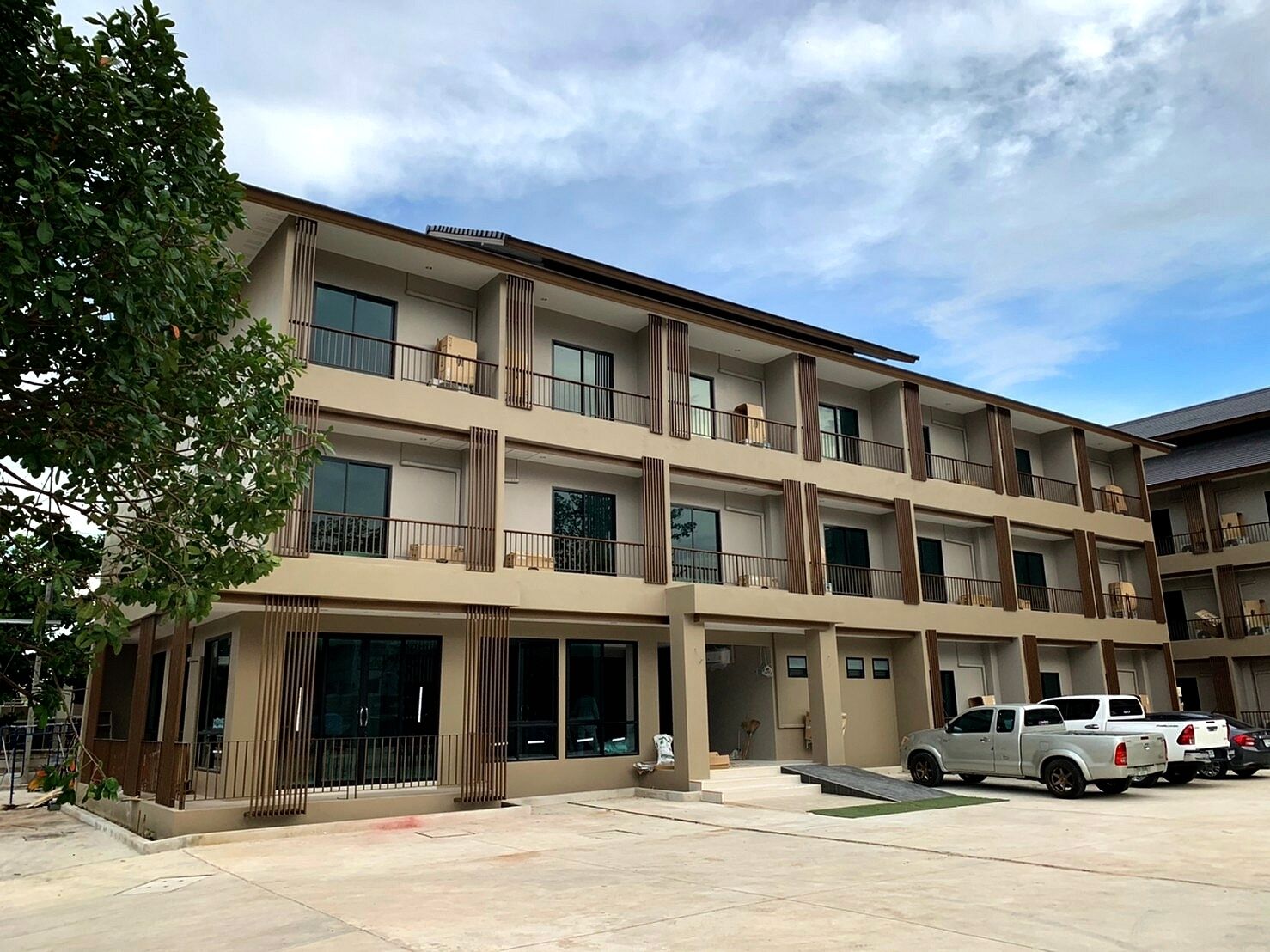 A multi-story residential building with balconies, large windows, and a parking area with vehicles.