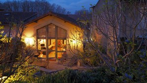 Ein gemütliches, helles Haus mit einer großen Glasdoor, umgeben von üppigem Grün und einem Ziegeldach.