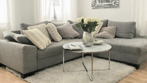 Una sala de estar moderna con un sofá gris, cojines blancos, una pequeña mesa redonda de centro y un jarrón con flores.