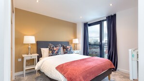 A hotel room with a bed, two bedside tables with lamps, a red throw blanket, and a view of the city through large windows. - Riverview Apartments (Glasgow)