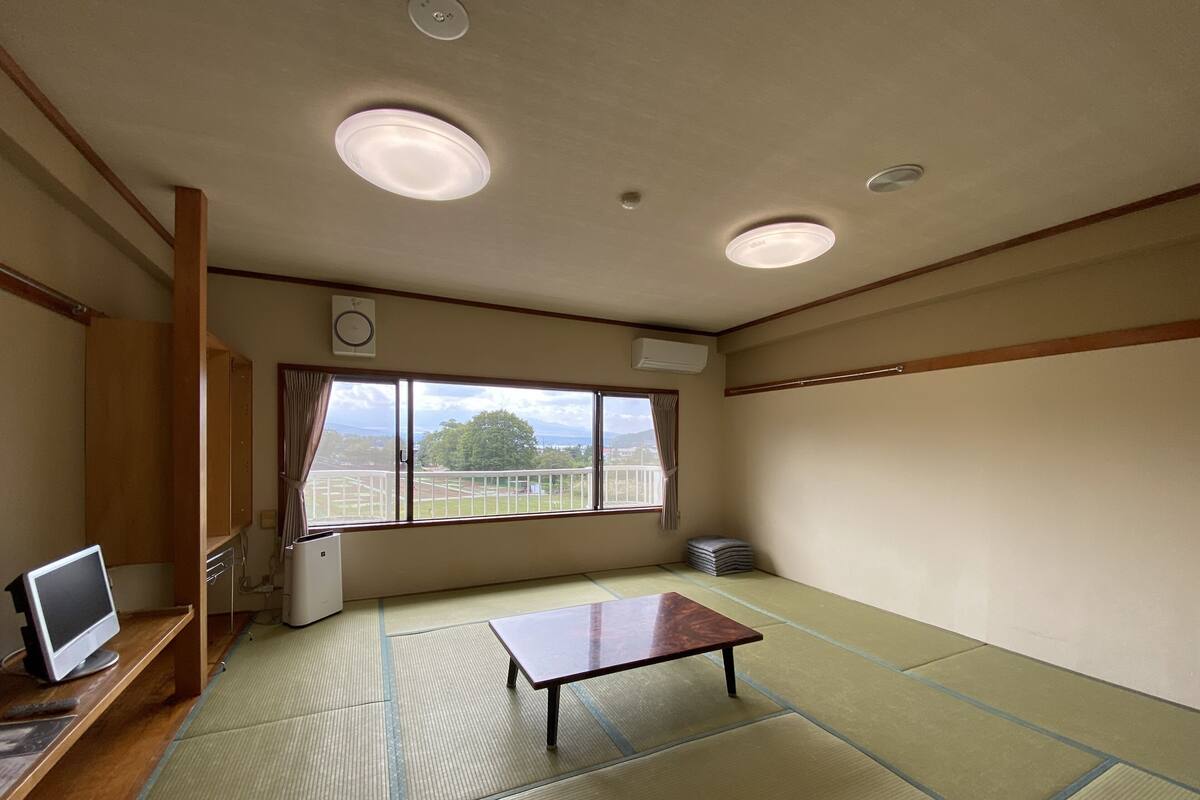 Une pièce dotée d’un sol recouvert de tatamis, d’une basse table en bois, d’un téléviseur posé sur un meuble et d’une fenêtre donnant sur un paysage verdoyant et des montagnes.