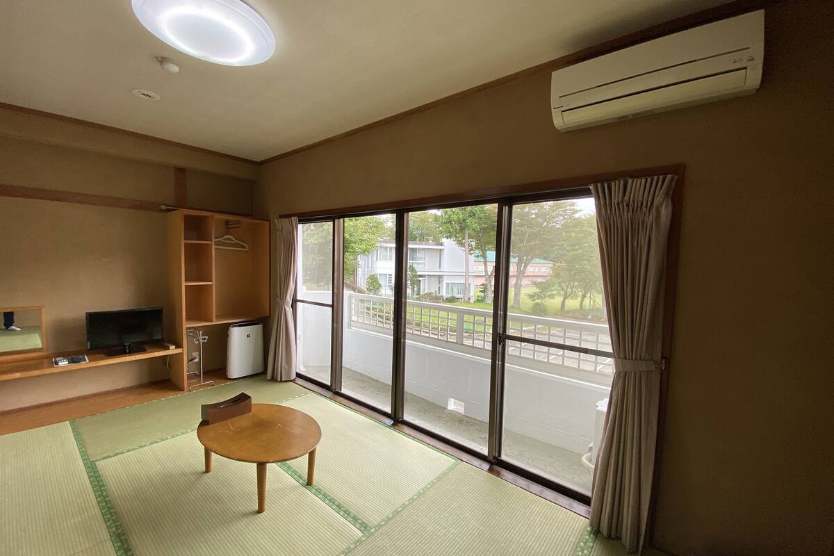 Une pièce avec un sol recouvert de tatamis, une table en bois, un téléviseur, une armoire et une grande fenêtre avec des rideaux.