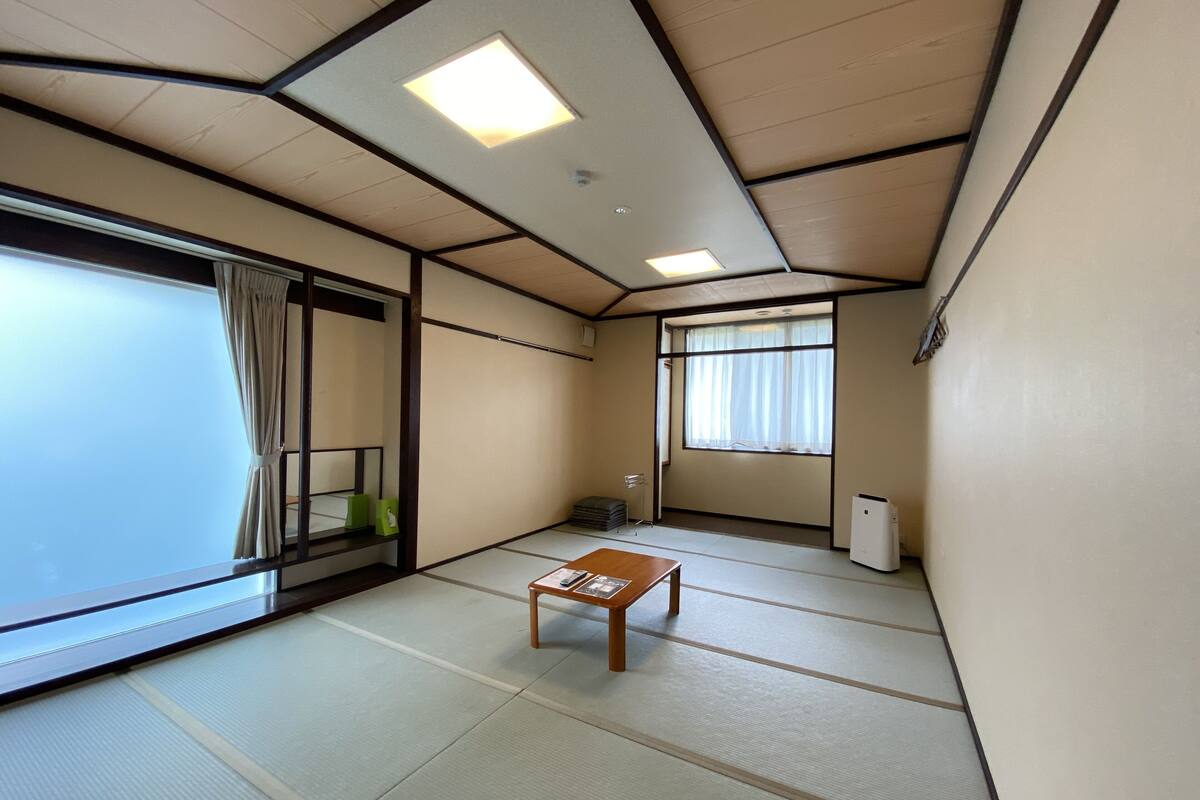 Une pièce de style japonais traditionnel, avec un sol recouvert de tatamis, une petite table en bois, des portes coulissantes et une grande fenêtre avec des rideaux.