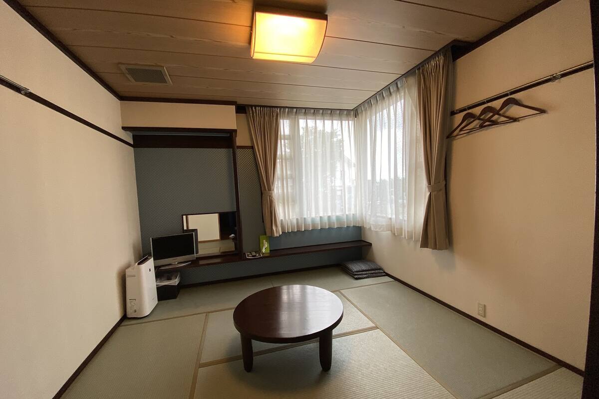 Une pièce de style japonais traditionnel, avec un sol recouvert de tatamis, une table basse, un téléviseur et une fenêtre ornée de rideaux légers.