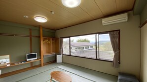 Een kamer met tatami vloerbedekking, een houten tafel, een televisie, een kledingkast en een raam met uitzicht op een huis.