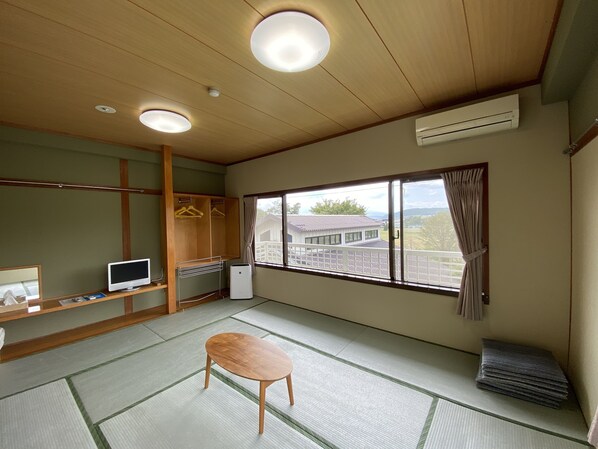 A room with tatami flooring, a wooden table, a TV, a wardrobe, and a window with a view of a house. - Green Hills New Minami (Yamanakako)