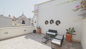 A rooftop terrace with a white wall, three circular wall decorations, a black metal bench, two potted cacti, and a view of buildings in the background. - Conte Orsini Apartment (Conversano)