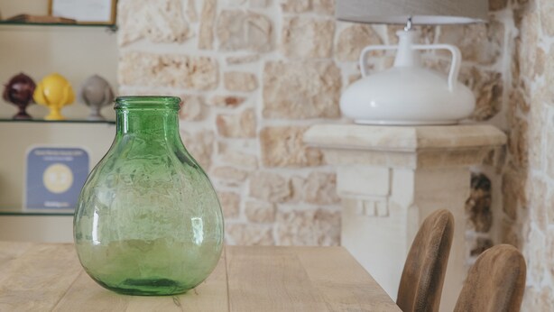 A dining table with a green glass vase, wooden chairs, and a stone wall background. - Conte Orsini Apartment (Conversano)
