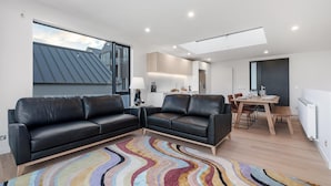 A modern living room with a black leather sofa, a colorful rug, and a dining area with a wooden table and chairs.