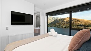 A bedroom with a large bed, a TV mounted on the wall, a bathroom visible through an open door, and a scenic mountain view through a large window.