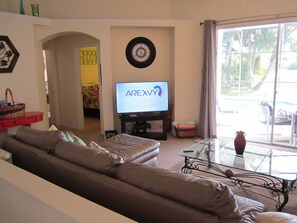 A living room with a brown leather sofa, a television displaying 'AREXYY', and a glass table with a vase.
