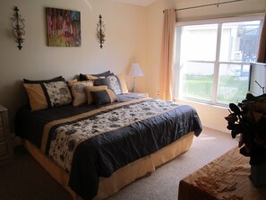A bedroom with a large bed, a bedside table, a lamp, a window with curtains, and a painting on the wall.
