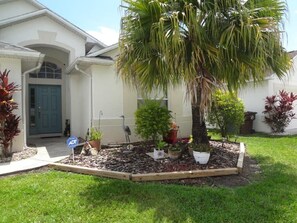 A residential house with a front yard featuring a palm tree, various potted plants, and a driveway.