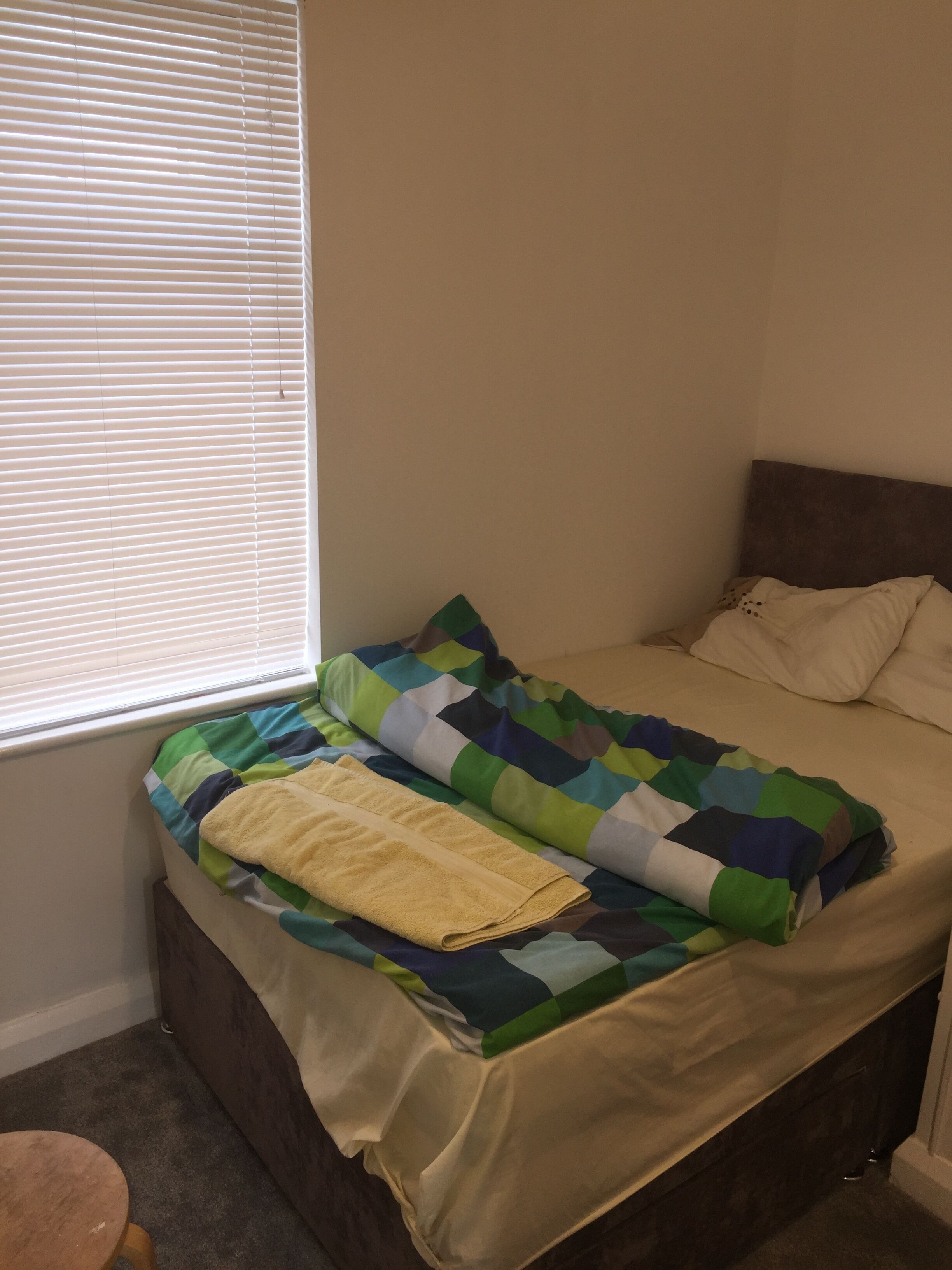 A bed with a multicolored quilt and a yellow towel, a window with blinds, and a wooden stool.