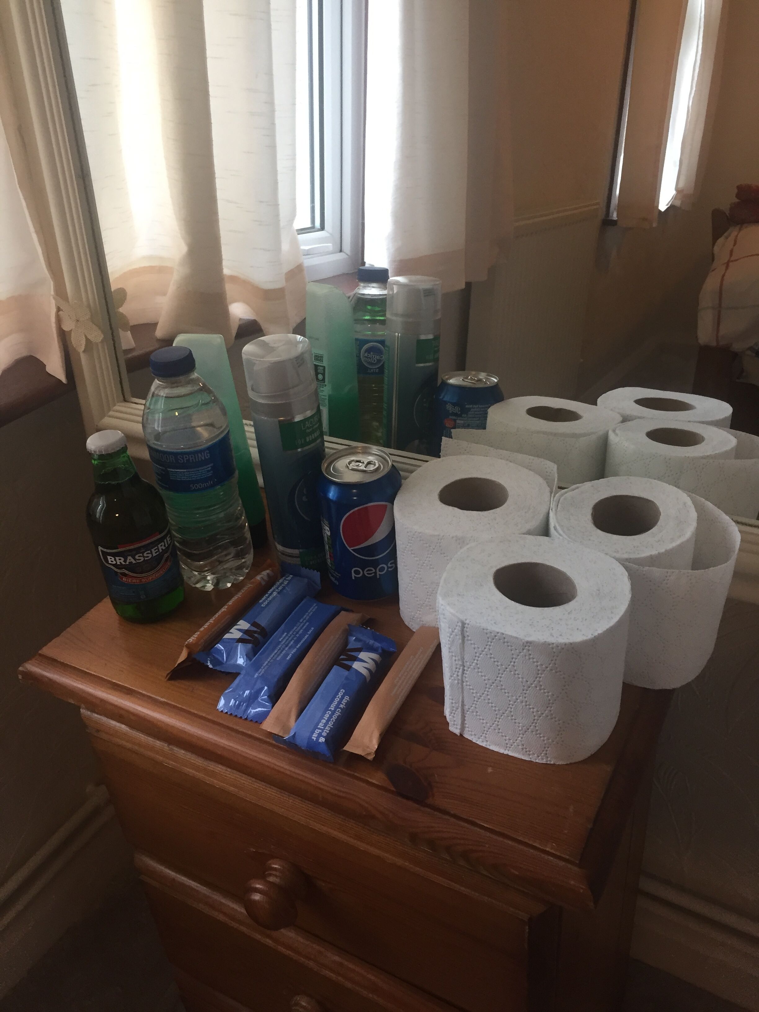 Une commode en bois contenant des articles de toilette, notamment des bouteilles d’eau, une canette de Pepsi et plusieurs rouleaux de papier toilette.