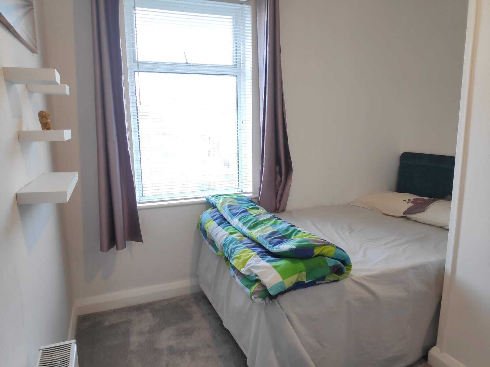 Een kleine, verzorgde slaapkamer met een enkel bed, een raam met gordijnen en een wandplank.