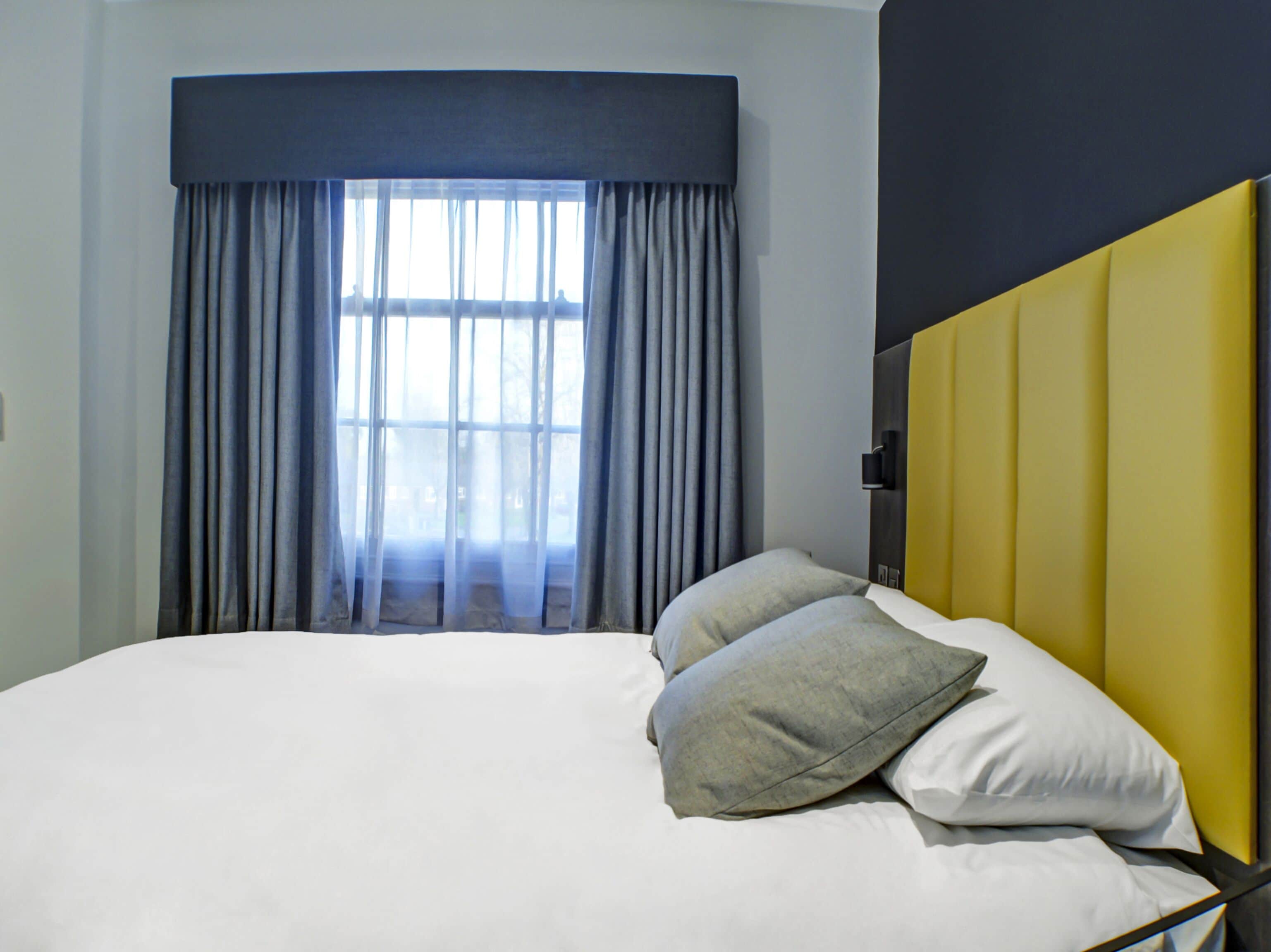 Una habitación de hotel moderna con una cama grande, un ventanal con cortinas y un cabecero amarillo.