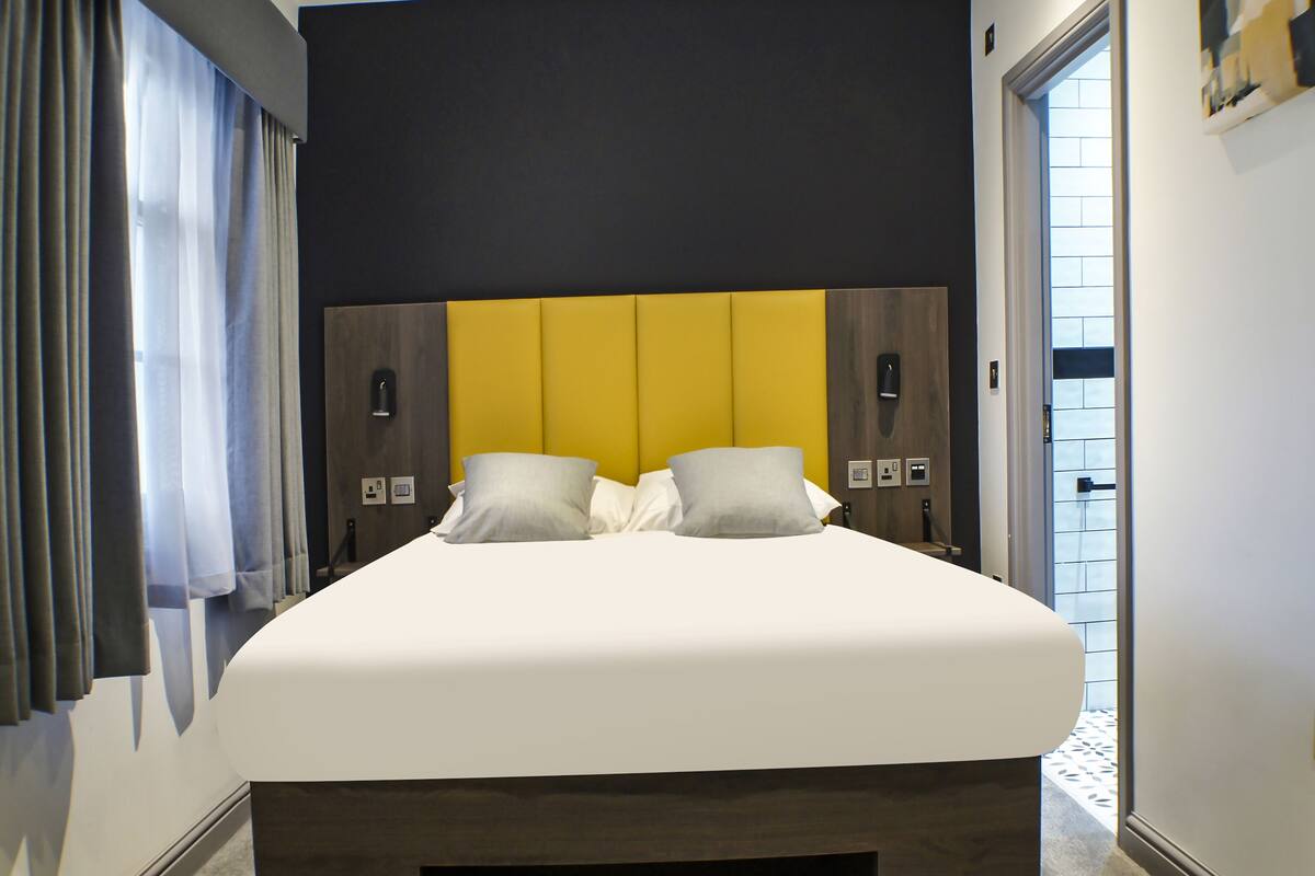 Ein modernes Hotelzimmer mit einem großen Bett, einer gelben Kopfteil und Blick ins Freie durch eine Tür.