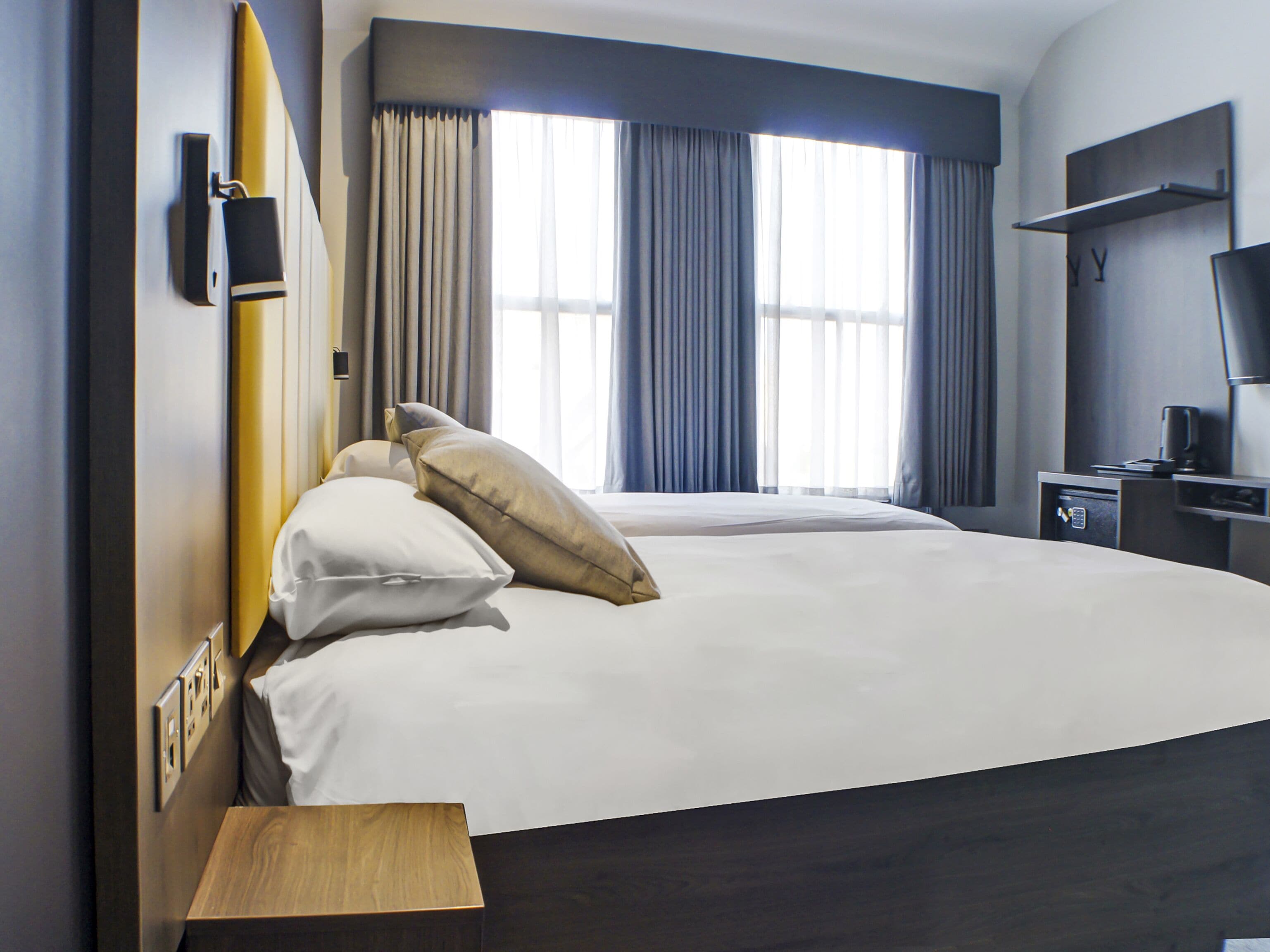 A hotel room with a large bed, two pillows, a bedside table, a nightstand, a TV, and a window with curtains.