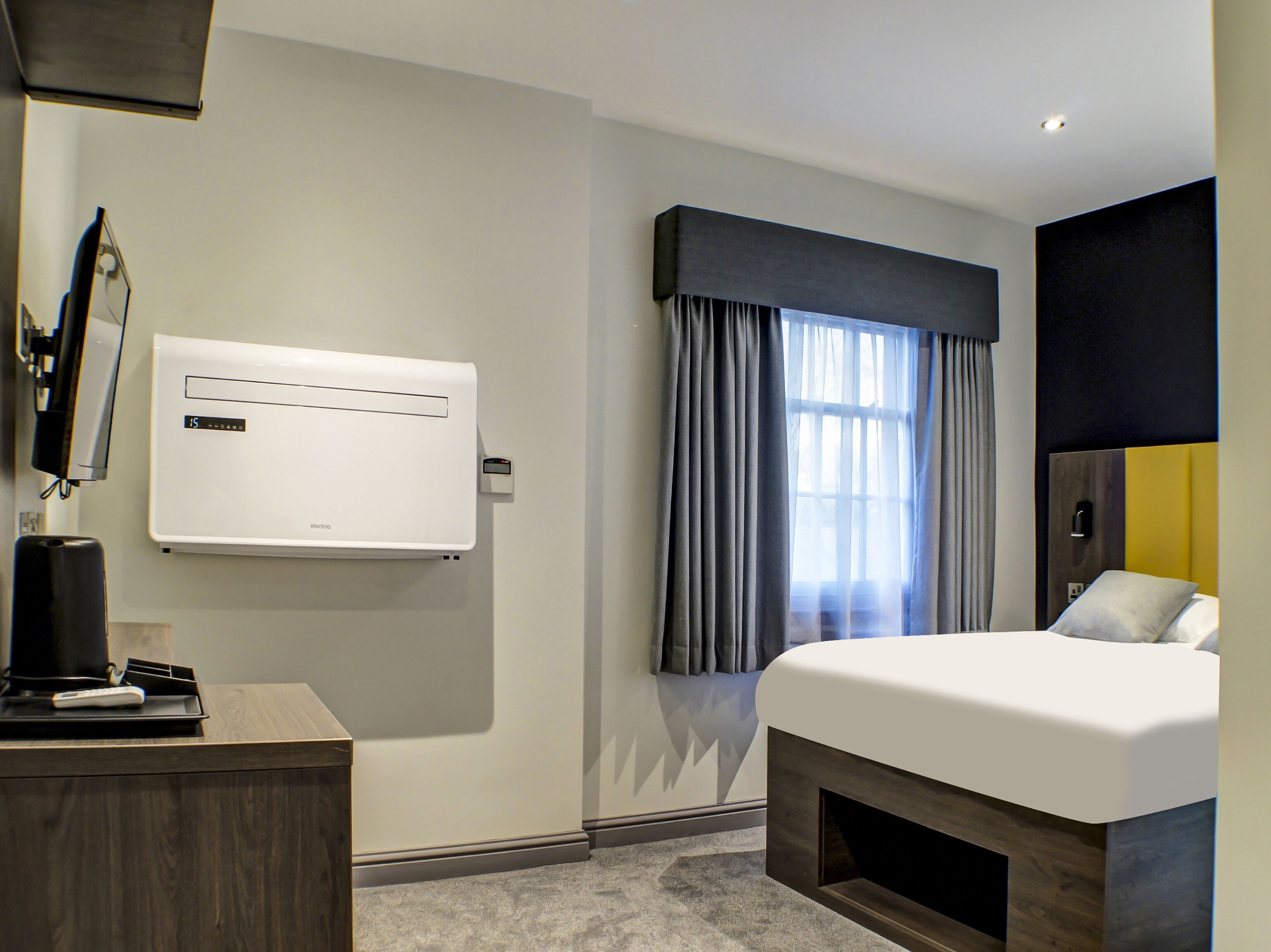 A hotel room with a bed, a desk with a coffee maker, an air conditioner, and a window with curtains.