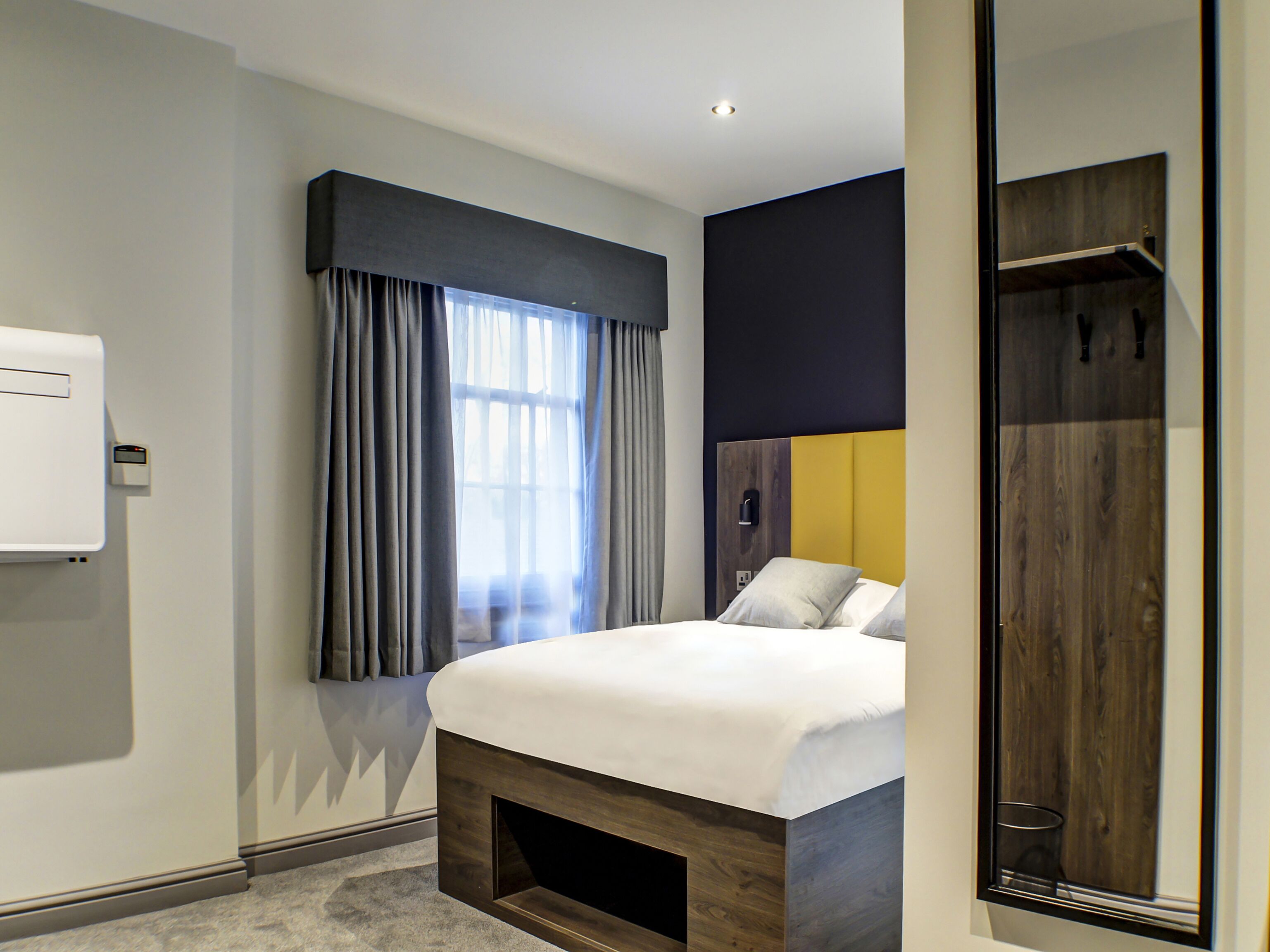 Una habitación de hotel moderna con cama, armario, espejo y ventana con cortinas.