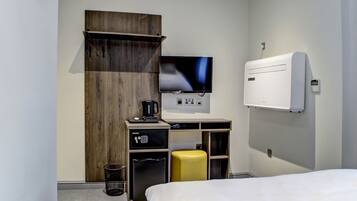 A modern hotel room with a bed, a desk with a computer, a TV, and a minibar.