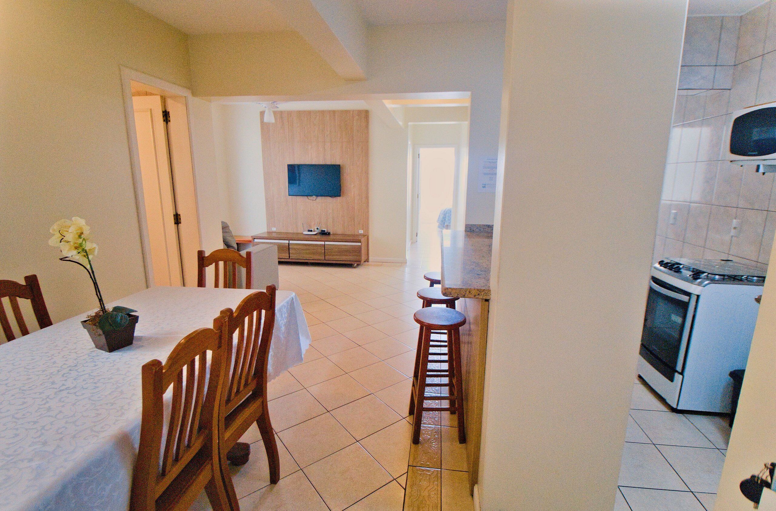 A dining area with a table, chairs, and a potted plant. There is a kitchen with a stove and a television mounted on the wall.