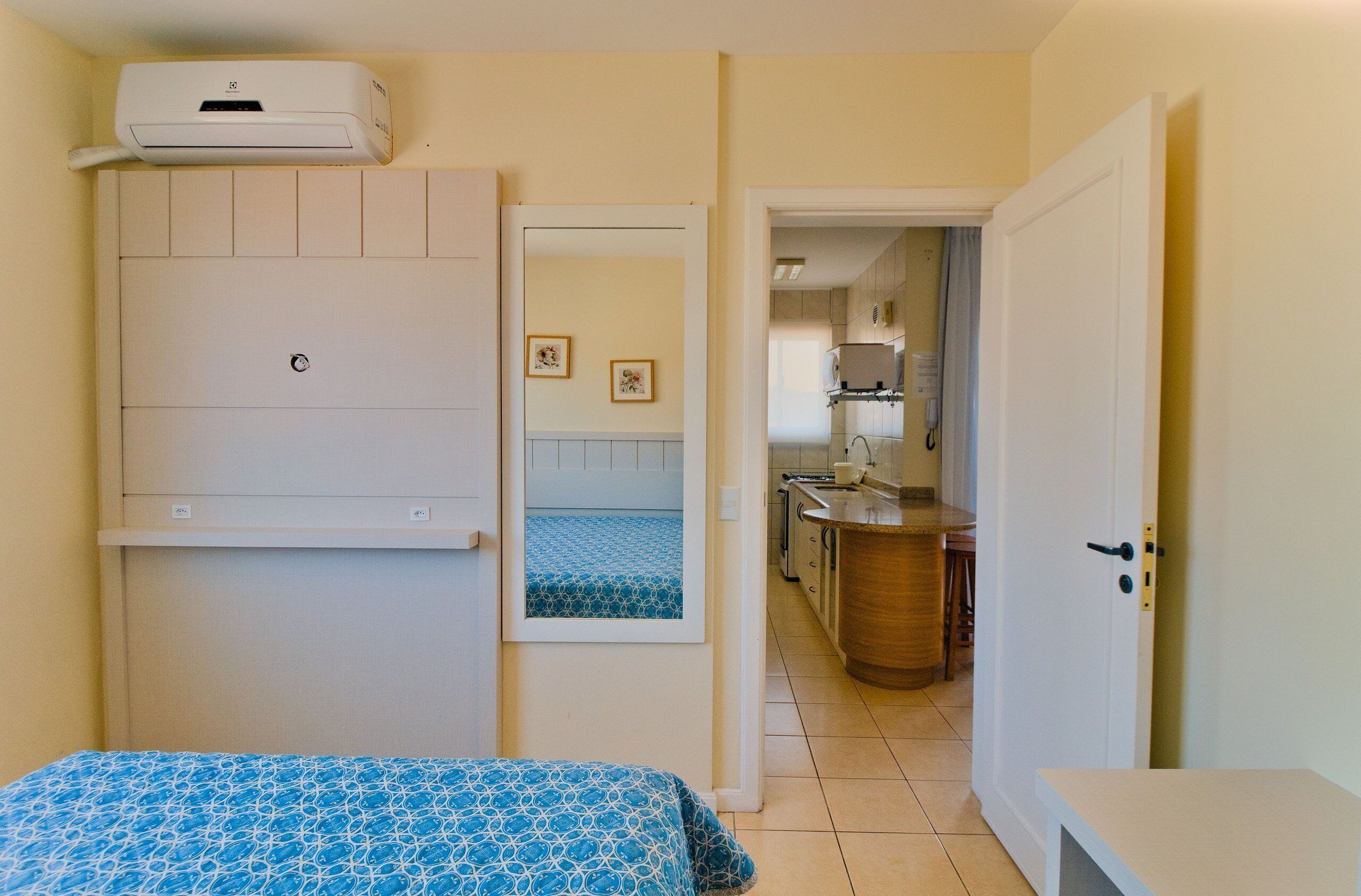 A room with a bed, a wall-mounted air conditioner, a small kitchen area, and a door leading to another room.