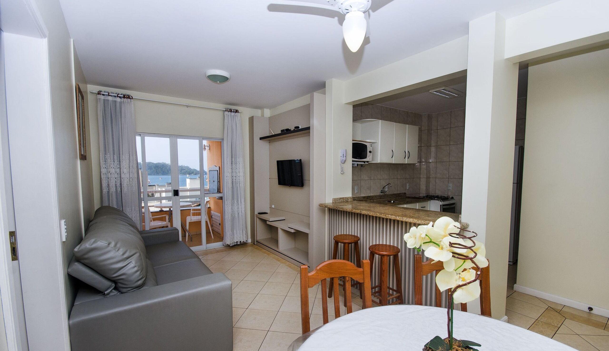 A modern apartment with a grey sofa, a dining table with chairs, a kitchen area, and a balcony with a view of the sea.